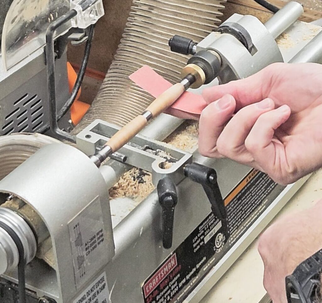 sanding the turned pen blank with sandpaper on a lathe while the lathe is running